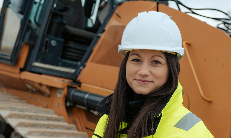 Female construction worker with white helmet and safety vest in front of a dozer. 